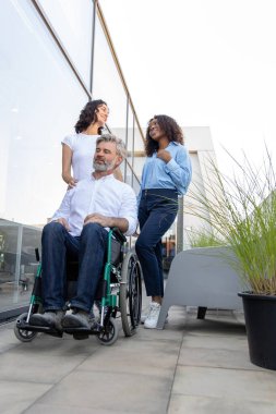 Ofisin yakınında. Ofis binasının yakınında tekerlekli sandalyede oturan bir adamla konuşan iki kadın.