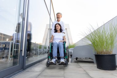 Şefkatli adam. Adam karısının tekerlekli sandalyesini taşıyor ve ona sevgiyle bakıyor.