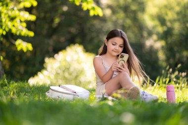 Parkta ağaçların yanında oturan, cep telefonu kullanan, interneti keşfeden ve dışarıda eğlenen sevimli bir çocuk.