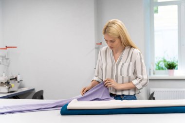 Tasarımcı. Sarışın, genç, beyaz elbise tasarımcısı kumaşla çalışıyor ve konsantre görünüyor.