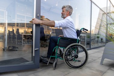 Ofis kapısında. Gri saçlı, sakallı, tekerlekli sandalyedeki olgun adam ofisin kapısını açıyor.