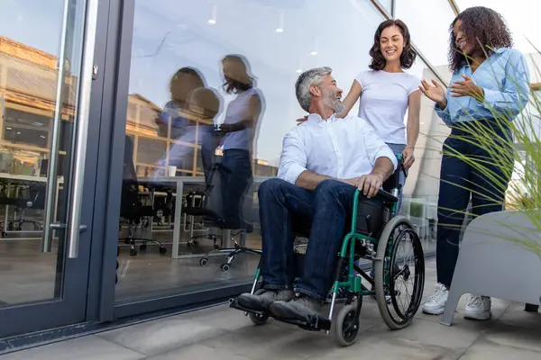 Ofisin yakınında. Ofis binasının yakınında tekerlekli sandalyede oturan bir adamla konuşan iki kadın.