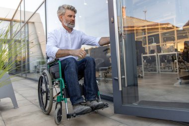 Engelli. Ofis bölgesinde tekerlekli sandalyede oturan gri saçlı, olgun bir adam.