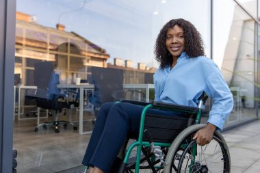 Ofisin yanındaki kadın. Koyu tenli, gülümseyen, engelli bir kadın ofis binasının yakınında tekerlekli sandalyede.