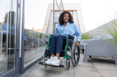 Ofisin yanındaki kadın. Koyu tenli, gülümseyen, engelli bir kadın ofis binasının yakınında tekerlekli sandalyede.