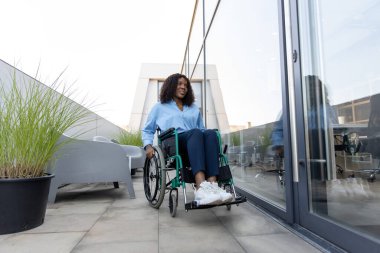 Ofisin yanındaki kadın. Koyu tenli, gülümseyen, engelli bir kadın ofis binasının yakınında tekerlekli sandalyede.