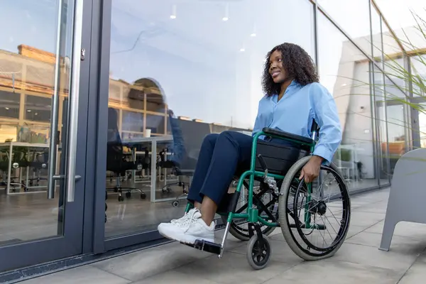 Ofisin yanındaki kadın. Koyu tenli, gülümseyen, engelli bir kadın ofis binasının yakınında tekerlekli sandalyede.