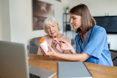 Genç dost canlısı hemşire, yaşlı kadın hasta ilaçlarını klinikte ya da bakımevinde dizüstü bilgisayarla açıklıyor.