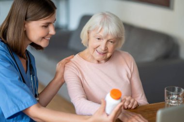Gülümseyen kadın doktor yaşlı hastaya ağrı kesici kullanma talimatlarını açıklıyor.
