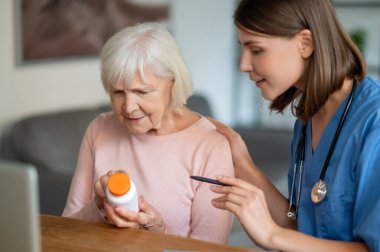 Genç dost canlısı hemşire klinikte ya da bakımevinde yaşlı kadın hasta ilaçlarını açıklıyor