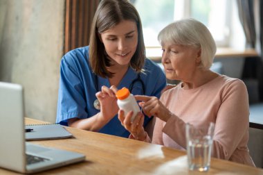 Genç dost canlısı hemşire klinikte ya da bakımevinde yaşlı kadın hasta ilaçlarını açıklıyor