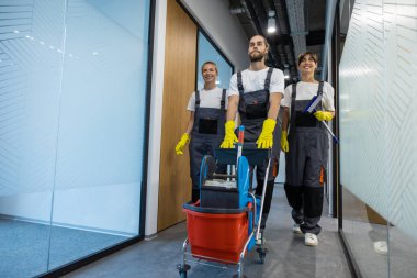 Profesyonel temizlikçiler. Temizlik ekibi temizlik malzemeleriyle ofis koridorunda yürüyor.
