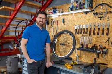 Mavi gömlekli tamirci atölyede alet ve bisiklet parçalarıyla poz veriyor.