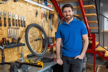 Mavi gömlekli sakallı tamirci atölyede aletlerle poz veriyor.