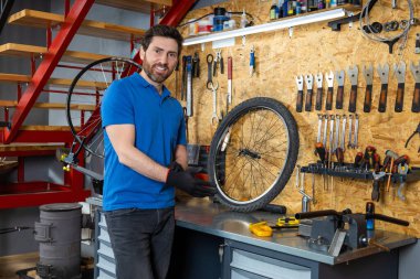 Mavi gömlekli sakallı tamirci atölyede aletlerle siyah eldiven giyiyor.