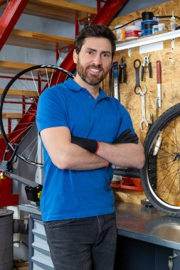 Mavi gömlekli tamirci atölyede kollarını kavuşturmuş aletlerin yanında duruyor.