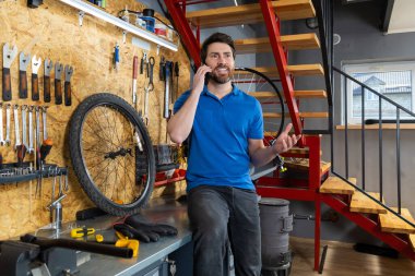 Mavi gömlekli tamirci atölyede bisiklet parçalarının yanında telefon ahizesiyle konuşuyor.