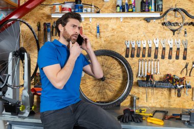 Mavi gömlekli tamirci atölyede bisiklet tekerinin yanında akıllı telefondan konuşuyor.
