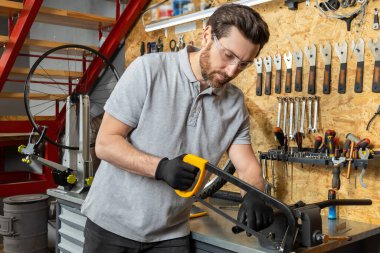 Demir testereli tamirci atölyede metal kesiyor ve arka planda tahta üzerinde aletler var.