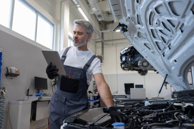 Otomobil tamir atölyesinde elinde tabletle tamirci açık kaputun yanında teşhis koyuyor.