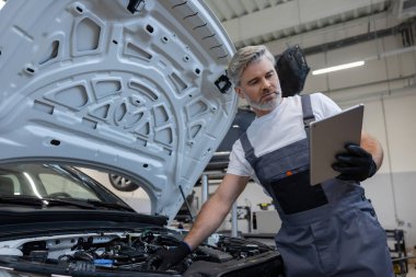 Tamirci otomobil tamirhanesinde motor kaputu açık tabletle teşhis koyuyor.