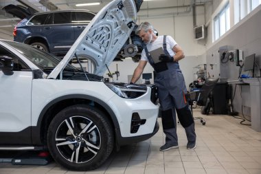 Otomobil tamir atölyesinde elinde tabletle tamirci açık kaputun yanında teşhis koyuyor.