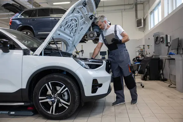 Otomobil tamir atölyesinde elinde tabletle tamirci açık kaputun yanında teşhis koyuyor.