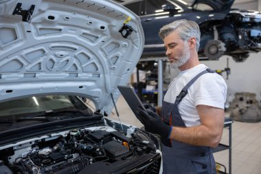 Otomobil tamir atölyesinde elinde tabletle tamirci açık kaputun yanında teşhis koyuyor.