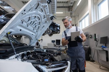 Tamirci otomobil tamirhanesinde motor kaputu açık tabletle teşhis koyuyor.