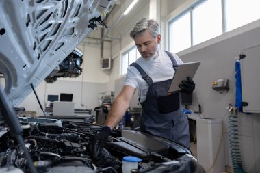 Oto tamir atölyesinde kaputu açık motor kontrolü için tablet kullanan tamirci