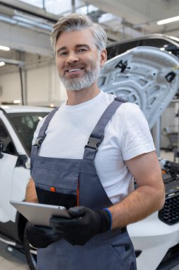 Araba tamirhanesinde kaputu açık bir şekilde duran tabletli ve eldivenli tamirci.