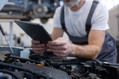 Otomobil tamir atölyesinde elinde tabletle tamirci açık kaputun yanında teşhis koyuyor.