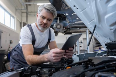 Otomobil tamir atölyesinde elinde tabletle tamirci açık kaputun yanında teşhis koyuyor.