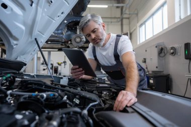 Tamirci otomobil tamirhanesinde motor kaputu açık tabletle teşhis koyuyor.