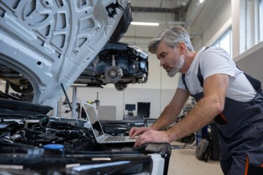 Motor kaputu açık olan oto tamir atölyesinde sistem kontrolü için dizüstü bilgisayar kullanan tamirci