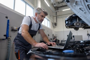 Oto tamir atölyesinde kaputu açık motor tanılama için dizüstü bilgisayar kullanan bir tamirci.