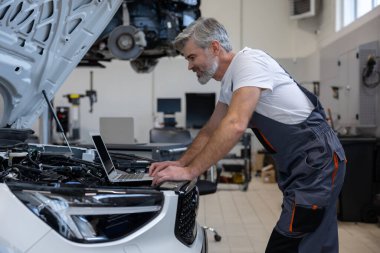 Tamirci, oto tamirhanesinde motor kaputunun altında dizüstü bilgisayarla teşhis koyuyor.