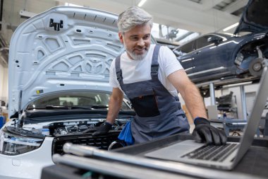 Tamirci, oto tamirhanesinde motor kaputunun altında dizüstü bilgisayarla teşhis koyuyor.