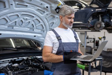 Tamirci, oto tamirhanesinde motor kaputunun altında dizüstü bilgisayarla teşhis koyuyor.