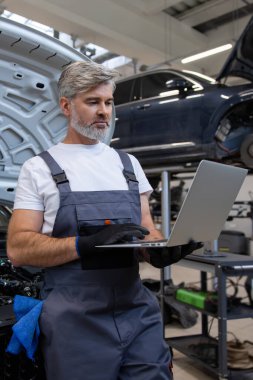 Tamirci, oto tamirhanesinde motor kaputunun altında dizüstü bilgisayarla teşhis koyuyor.