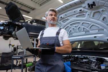 Motor kaputu açık olan oto tamir atölyesinde sistem kontrolü için dizüstü bilgisayar kullanan tamirci