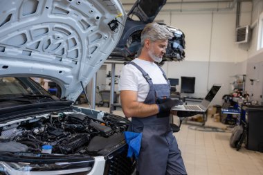 Motor kaputu açık olan oto tamir atölyesinde sistem kontrolü için dizüstü bilgisayar kullanan tamirci
