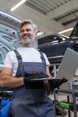Oto tamir atölyesinde kaputu açık motor tanılama için dizüstü bilgisayar kullanan bir tamirci.