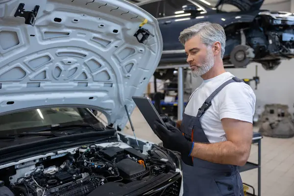 Otomobil tamir atölyesinde elinde tabletle tamirci açık kaputun yanında teşhis koyuyor.