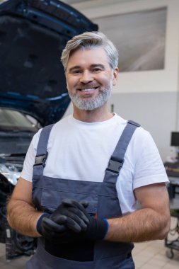 Tamirci arka planda kaputu açık bir şekilde oto tamirhanesinde dururken eldiven tutuyor.