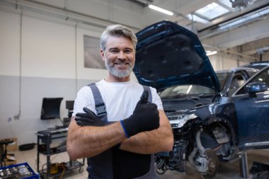 Tamirci, arka kaputu açık bir şekilde oto tamir atölyesinde dikilirken jest gibi duran eldivenler tutuyor.