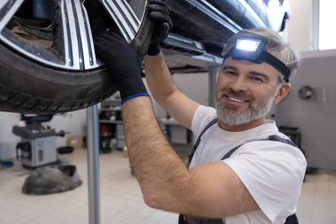 Tamirci çömelme sırasında oto tamirhanesinde araç çarkını kontrol ediyor