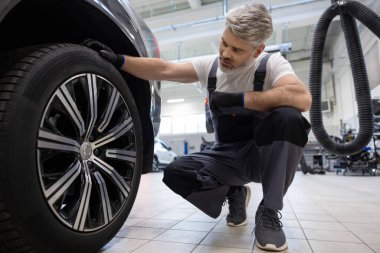 Tamirci çömelme sırasında oto tamirhanesinde araç çarkını kontrol ediyor