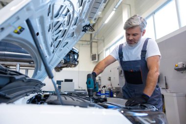 Tamirci oto tamirhanesinde kaputu açık motorda matkap kullanıyor.