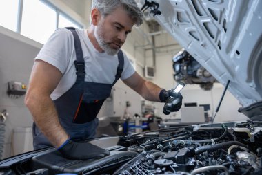 Otomobil tamirhanesinde kaputun altında el feneriyle motoru denetleyen tamirci.